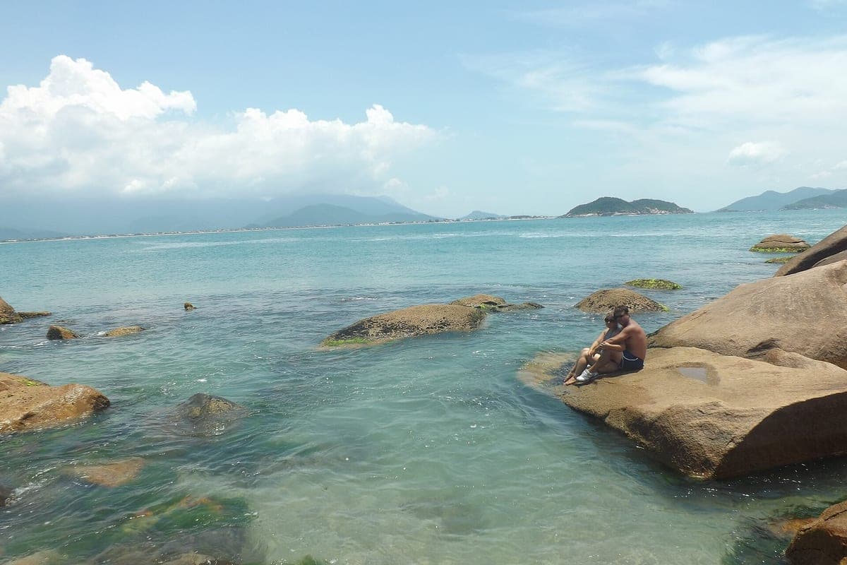 Água transparente sobre as pedras na Ponta da Pinheira, com uma pessoa sentada na rocha e ilhas no horizonte