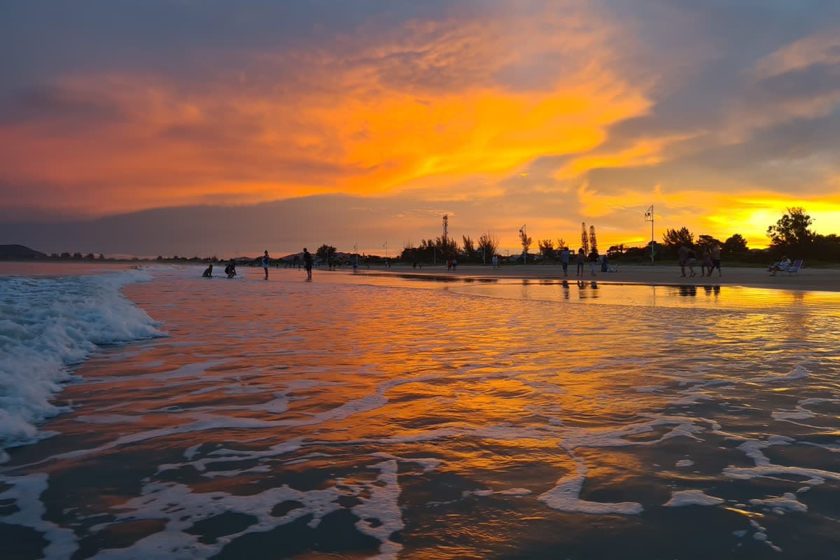 Pôr do sol na Praia do Sonho, com o céu alaranjado refletido na areia molhada e pessoas caminhando na beira